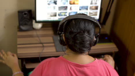 Indian-asian-caucasian-woman-girl-in-pink-t-shirt-listening-enjoying-music-wearing-headphone-on-desktop-pc-computer-at-home