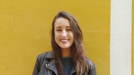 Slow-motion-portrait-of-a-fashionable,-young-woman-smiling-brightly-in-front-of-a-yellow-wall