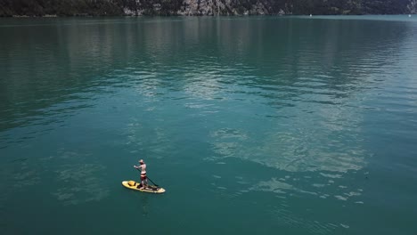 Drone,-aerial-orbiting-shot-around-a-male-stand-up-paddler-on-a-yellow-sup,-stand-up-paddle,-he-wears-a-straw-hat-in-the-middle-of-a-lake-in-Switzerland