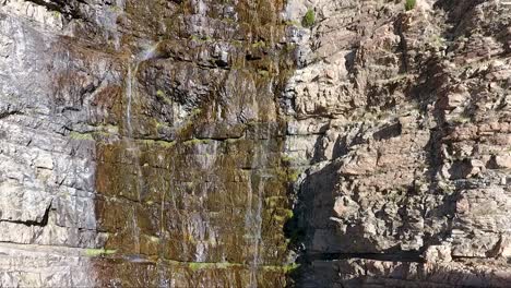 A-small-waterfall-in-Ogden-Canyon,-Utah-trickles-down-craggy-rocks-as-seen-by-an-ascending-drone-from-close-proximity