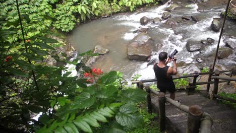 A-quick-pan-left-to-right-shot-from-wet,-damp-leaves,-to-a-young-white-man-in-black-clothing,-standing-taking-photos-of-a-river-and-a-waterfall-below-a-steep-set-of-stairs-in-the-jungle-of-Bali