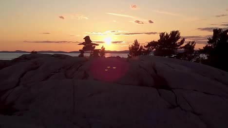 Reveal-of-Inukshuk-on-Rocky-Pine-Tree-Island-at-Sunset,-Drone-Aerial-Wide-Dolly-Out