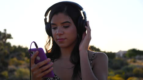 A-beautiful-young-hispanic-woman-holding-a-smartphone-listening-to-music-on-headphones-outdoors-in-epic-sunlight-with-lens-flares-in-slow-motion