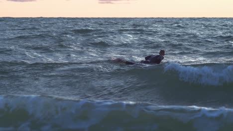 Surfer-on-surfboard-paddling-over-waves-near-the-Baltic-sea-Karosta-beach-at-Liepaja-during-a-beautiful-vibrant-sunset-at-golden-hour,-medium-shot-from-a-distance