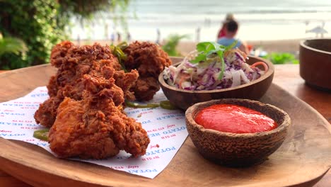 crispy-deep-fried-cricken-with-sriracha-hot-suace-and-coleslaw-on-wooden-cutting-board-with-sea-view,-filmed-in-bali-indonesia,-holiday-food