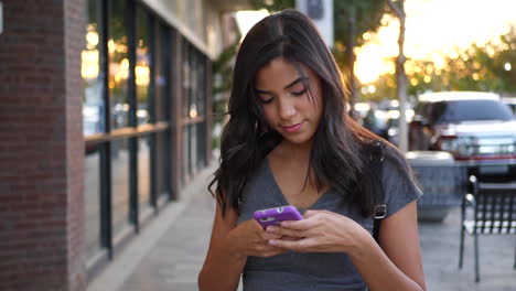 Eine-Attraktive-Junge-Hispanische-Frau,-Die-Durch-Die-Straßen-Der-Stadt-Geht,-SMS-Schreibt-Und-Glücklich-Auf-Eine-Nachricht-Auf-Ihrem-Handy-In-Zeitlupe-Schaut