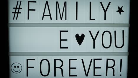 A-hand-completing-a-text-message-on-a-backlit-billboard:-Family-Love-You-Forever,-a-hashtag-and-some-symbols-