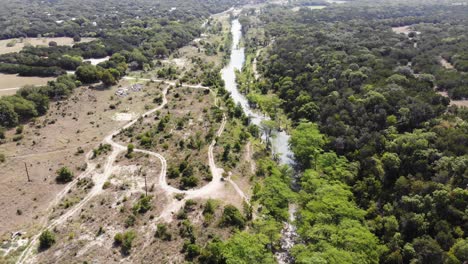 starting-above-area-of-river-heavily-shaded-by-cypress-trees,-flying-towards-large-field-with-roads-on-the-left---huge-expanse-of-trees-on-the-right---Aerial-footage-of-Blanco-river---Wimberly,-TX
