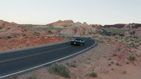 Black-Ferrari-convertible-sports-car-driving-along-on-a-highway-in-the-Valley-of-Fire,-Nevada,-USA,-at-sunset