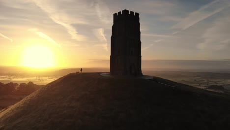 Zooming-out-aerial-footage-of-the-golden-sunrise-over-Glastonbury-Tor,-Somerset