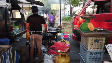 2-men-cooking-food-on-3-large-wok-with-red-truck,-gas-tank,-a-weighing-scale-and-plastic-containers-around-them