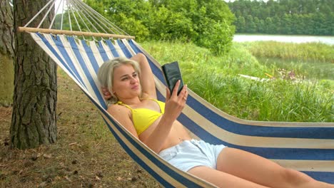 Blondes-Mädchen-Liegt-An-Einem-Warmen,-Sonnigen-Sommertag-In-Einer-Hängematte-Und-Führt-Einen-Videoanruf-Per-Handy