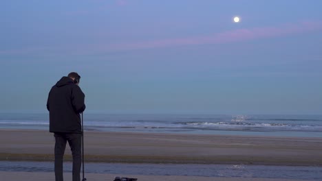 The-sound-technician-is-recording-noises-on-the-beach-at-sunset,-the-pink-sky-and-the-waves-of-the-sea-with-the-horizon-in-the-background-of-the-sequence