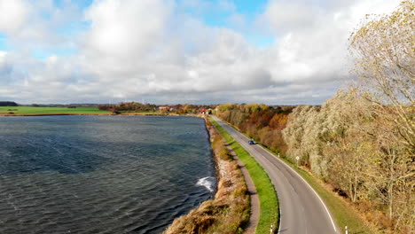 Car-driving-on-road-between-Schlei-sea-and-forest-in-autumn,-aerial-panning-shot