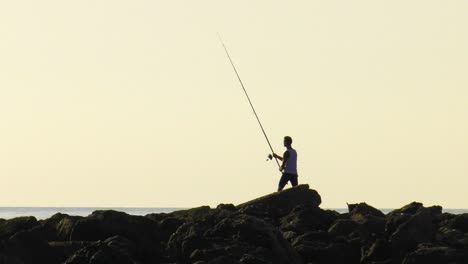 Young-man-casting-fishing-rod-from-rocks-at-dawn,-slow-motion-silhouette