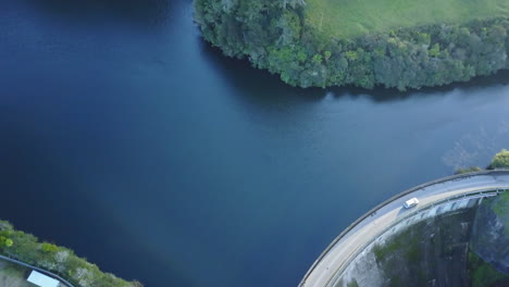 Aerial-shot-of-Car-driving-across-a-dam-with-blue-water-and-green-forest