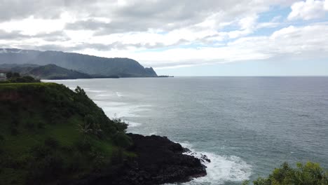 4K-Hawaii-Kauai-Tilt-down-from-mountains-in-distance-to-reveal-waves-crashing-on-rocky-shore-with-greenery-in-foreground