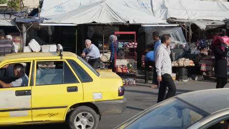 Cars-passing-on-the-road,-several-people-next-to-a-marketplace-in-Homs-City,-Syria