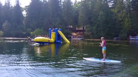 aerial-dolly-backwards,-a-boy-watches-from-a-floating-paddleboard-on-a-lake-as-other-kids-jump-off-an-inflatable-"blob"-toy-and-launch-each-other-into-the-air
