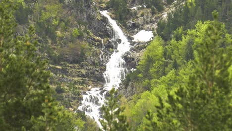 High-cascading-waterfalls-on-rocky-mountainside-seen-through-pine-trees,-zoom