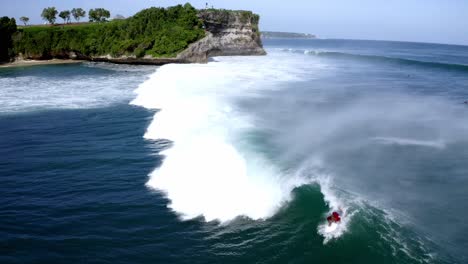 Male-surfer-in-red-shirt-catching-a-large-four-meter-wave-in-Balangan-Beach-while-its-crashing,-Aerial-dolly-out-tracking-shot