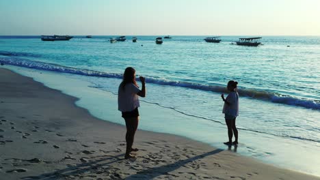 Touristin-Fotografiert-Einen-Freund-Am-Strand-Bei-Sonnenuntergang,-Umgeben-Von-Strahlend-Blauem-Meer-Und-Segelbooten-–-Weitwinkelaufnahme
