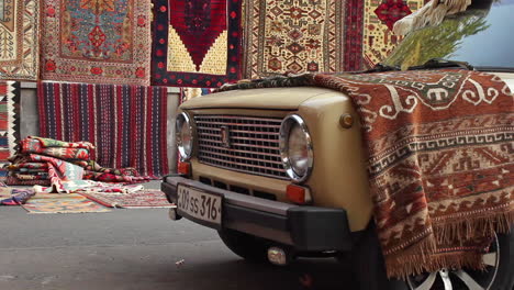 Parked-car-with-colourful-rug-draped-over-bonnet-against-background-of-hanging-handmade-rug