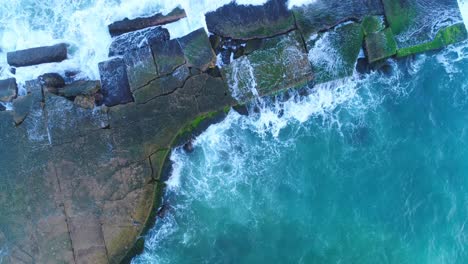 Scenic-view-of-stone-rocky-jetty-in-blue-green-ocean-sea-water-and-waves-crashing-onshore,-above-rising-aerial