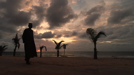 Ein-In-Afrikanisches-Dashiki-Gekleideter-Mann-Geht-Bei-Sonnenuntergang-Von-Links-Nach-Rechts-Am-Strand-Von-Senegambia,-Serekunda,-Gambia-Vorbei