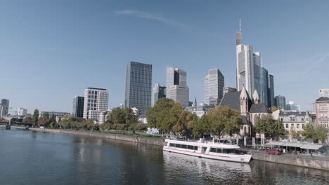 Static-shot-of-Frankfurt-Skyline-with-Skyscrapers-and-ferry-in-forefront,-view-from-main-river-on-a-sunny-day-with-clear-sky,-hessen,-Germany