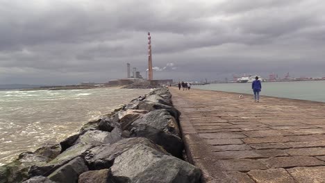 Some-waves-on-the-left-and-Dublin-ESB-Poolbeg-in-the-centre-with-a-shop-on-the-right,-some-people-walking-along-the-coast-or-south-wall