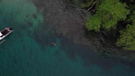 Aerial-shot-of-a-young,-white-man-is-swimming-with-fins-in-the-water-on-the-surface,-above-a-coral-reef-next-to-his-small-white-boat