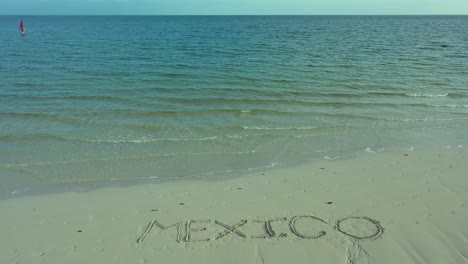 Wide-angle-shot-of-MEXICO-inscribed-in-the-sand-on-a-beach-with-a-windsurfer-on-the-horizon