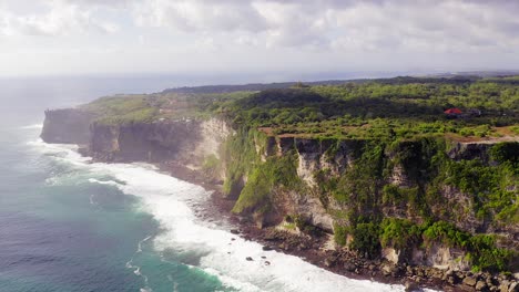 Uluwatu-cliffs,-Pull-back-aerial-view-revealing-the-sharp-edge-Cliff-with-lush-green-landscape,-tertiary-limestone-layers-and-Indian-Ocean-waves-breaking-on-the-beach,-Bali,-Indonesia