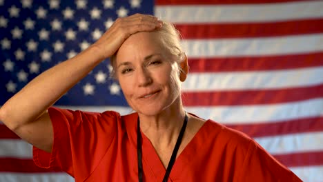 Medium-tight-portrait-of-nurse-looking-at-the-camera,-relieved-and-very-happy-with-a-US-flag-behind-her