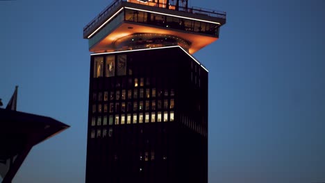 The-Iconic-A'dam-Tower-Lookout-In-Amsterdam,-Netherlands-With-Ferry-Boat-Cruising-On-A-Sunset---tilt-down-shot