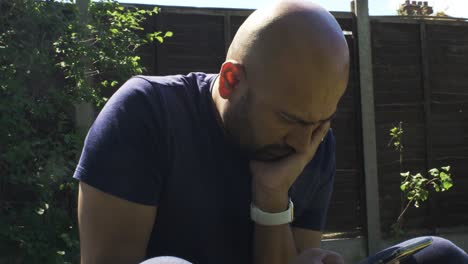 Male-Sitting-In-Garden-With-Face-Resting-On-Hand-Looking-Pensively-At-Phone