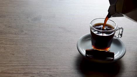 Gießen-Sie-Kaffee-In-Eine-Leere-Transparente-Tasse,-Unterstützt-Von-Einer-Weißen-Untertasse-Auf-Einem-Holztisch