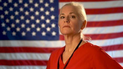 Medium-tight-portrait-of-the-back-of-blonde-nurses-head-as-she-turns-and-looks-at-camera-looking-angry-and-concerned-with-out-of-focus-US-flag
