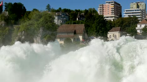 Schwenk-Eines-Riesigen-Wasserfalls-Und-Städtischer-Gebäude-Im-Hintergrund-An-Einem-Sonnigen-Tag-Mit-Blauem-Himmel,-Rheinfall-In-Der-Schweiz