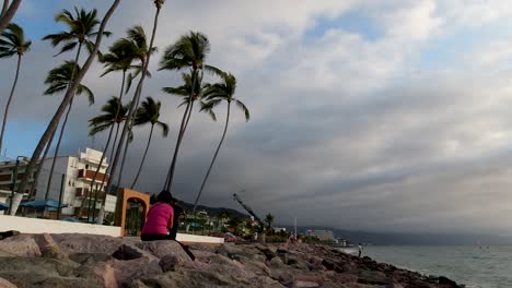 A-person-sitting-on-rocks-during-sunset-next-to-the-sea-in-Puerto-Vallarta,-Mexico