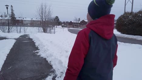 Young-child-walking-on-cleared-path-in-winter-wearing-red-and-white-jacket-and-blue-toque-with-a-pompom