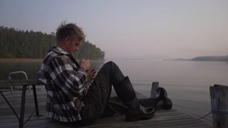 A-man-sits-down-on-a-jetty-in-a-lake-to-drink-out-of-his-cup-in-the-morning
