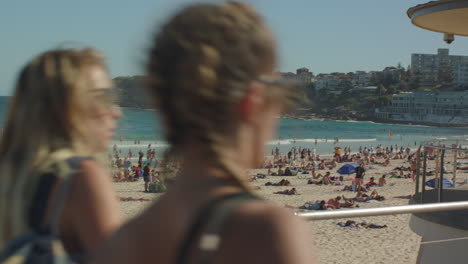 Ein-Belebter-Bondi-Strand-Mit-Ein-Paar-Frauen,-Die-Die-Kamera-Kreuzen-Und-Das-Haus-Der-Lebensretter-In-Sydney,-Australien,-Enthüllen