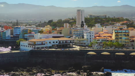 Turistas-Caminando-En-El-Puerto-De-Puerto-De-La-Cruz,-Tenerife,-Islas-Canarias,-Sus-Muelles-De-Piedra,-Rocas-Y-Arrecifes-Bañados-Por-Las-Olas-Del-Océano-Atlántico-En-Un-Día-Soleado-De-Verano,-Toma-Aérea-4k