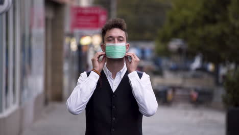 An-elegant-young-man-wearing-a-black-waistcoat-and-a-white-shirt-putting-on-a-green-protective-Covid-19-mask,-standing-on-a-busy-street,-looking-straight-into-the-camera,-static-close-up-4k-shot