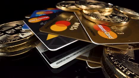 Close-up-turning-Bitcoins-BTC-and-MasterCard-credit-debit-cards-on-black-reflective-glass-surface,-4k-cryptocurrency-payments