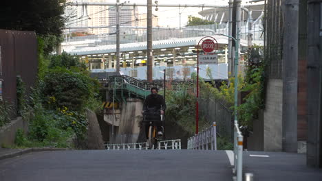 Hombre-Asiático-Con-Mascarilla-En-Bicicleta-Cuesta-Arriba-En-La-Calle-De-Tokio,-Japón-Durante-La-Pandemia