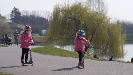 Adorable-Children,-Twin-Sisters-With-Face-Masks-Riding-Scooters-in-Park-by-Lake-During-Covid-19-Virus-Pandemic