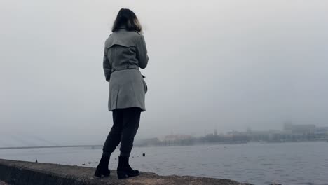 Girl-standing-near-misty-river-and-looking-how-city-disappears,-meanwhile-vaping